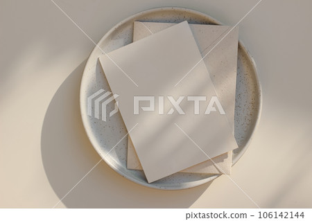 Summer minimal wedding stationery. Blank vertical greeting card mockup with beige envelope, invitation. Ceramic dessert plate in golden sunlight. Table background with shadows overlay. Flatlay, top 106142144