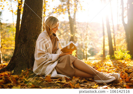 Beautiful young woman sitting on a fallen autumn leaves in a park, reading a book. Relaxation. 106142516