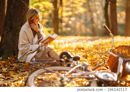 Beautiful young woman sitting on a fallen autumn leaves in a park, reading a book. Relaxation. 106142517