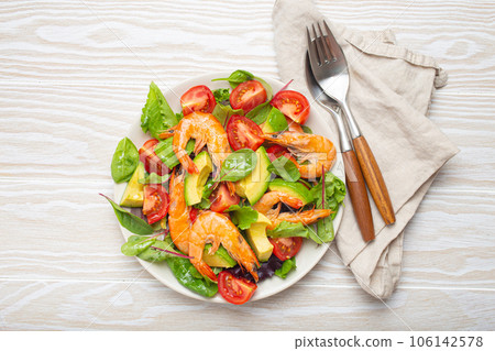 Healthy salad with grilled shrimps, avocado, cherry tomatoes and green leaves on white plate with cutlery on white wooden rustic background top view. Clean eating, nutrition and dieting concept. 106142578