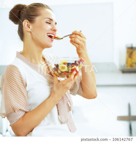 Happy young housewife eating fresh fruit salad in kitchen Happy young housewife eating fresh fruit salad in kitchen 106142767