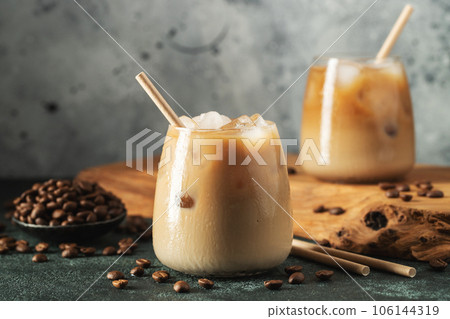 Ice coffee in a tall glass with cream poured over, ice cubes and beans on a dark concrete table. 106144319