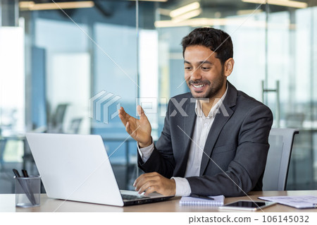 Young successful arab businessman inside office at workplace talking remotely with colleagues and partners, man using laptop for video call, satisfied boss smiling and gesturing with hands joyfully. 106145032