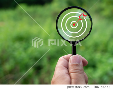 Human hand holding a magnifying glass with target icon on green nature background. Business concept. Management concept. Business planning concept. Human hand holding a magnifying glass with target icon on green nature background. Business concept. Management concept. Business planning concept. 106146432