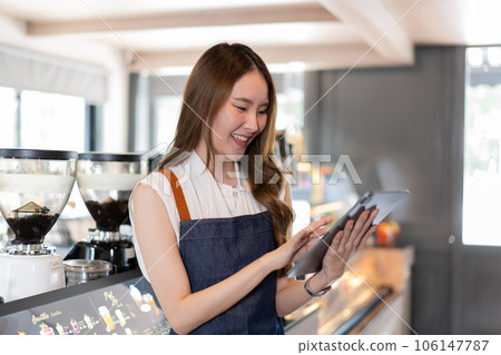Young Asian female coffee shop owner and barista using a digital tablet. Millennial Asia woman smiling and working at cafe and restaurant indoors. Technology usage Young Asian female coffee shop owner and barista using a digital tablet. Millennial Asia woman smiling and working at cafe and restaurant indoors. Technology usage 106147787