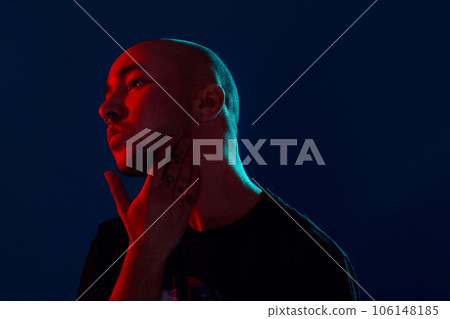 Studio shot of a young tattoed bald man posing against a blue background. 90s style. Studio shot of a young tattoed bald man posing against a blue background. 90s style. 106148185