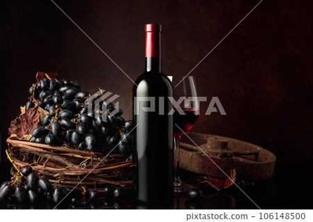 Glass and bottle of red wine with blue grapes on a black reflective background. 106148500
