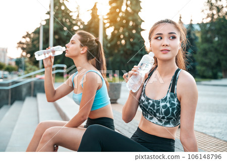Athletic young women relaxing and drinking water sitting on the steps after workout in urban environment 106148796