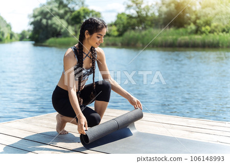 Beautiful Indian girl laying yoga mat on pier by the water. Outdoors yoga training and healthy lifestyle. Beautiful Indian girl laying yoga mat on pier by the water. Outdoors yoga training and healthy lifestyle. 106148993