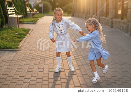Two little sisters are happily spinning on the city streets at sunset. Funny girls laugh holding hands. Two little sisters are happily spinning on the city streets at sunset. Funny girls laugh holding hands. 106149128