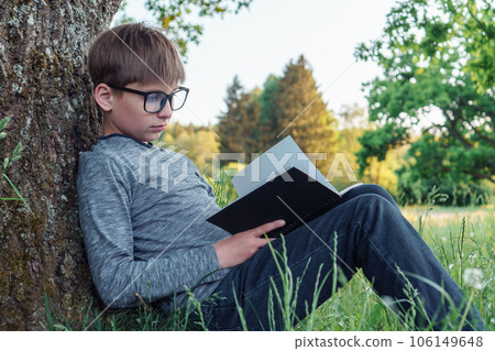 Mindful boy in glasses sitting on green lawn leaning on tree in park and reading task in notebook, studying outdoors. 106149648