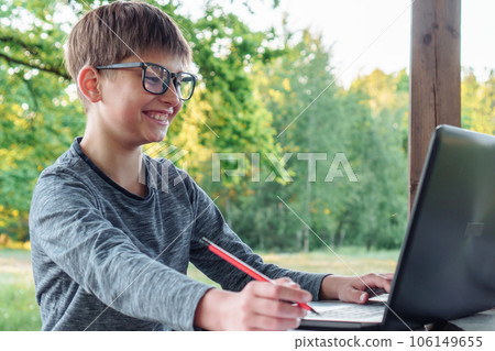 Boy with glasses sit at park table, looking at laptop screen and smiling. Pupil holding pencil, enjoy online studying. Boy with glasses sit at park table, looking at laptop screen and smiling. Pupil holding pencil, enjoy online studying. 106149655