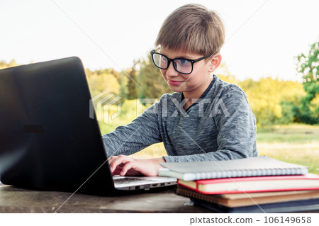 Junior student in glasses smiling while study with laptop in park. Boy staring at screen of gadget with attentive eyes. 106149658