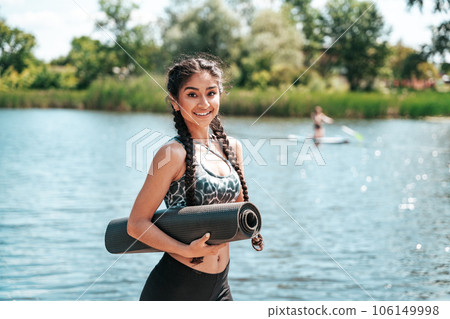 Beautiful Indian woman with a fitness mat in her hands with a river in the background getting ready to do yoga 106149998