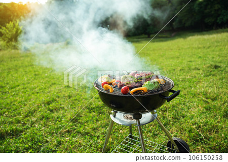 Grilled meat in grill. Roasting steaks and vegetables bbq in a round grill in a meadow. Summer picnic outdoors. Grilled meat in grill. Roasting steaks and vegetables bbq in a round grill in a meadow. Summer picnic outdoors. 106150258