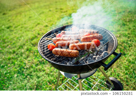 Grilled sausages grill. Roasting sausages bbq with tomatoes and rosemary in a round grill in a meadow. Summer picnic outdoors. Grilled sausages grill. Roasting sausages bbq with tomatoes and rosemary in a round grill in a meadow. Summer picnic outdoors. 106150281