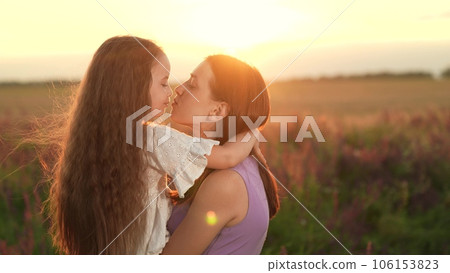 Mother and little daughter kiss observing bright sunset on country field. Pretty daughter and mother spend time in wild field. Careful mother embraces daughter standing in evening park field Mother and little daughter kiss observing bright sunset on country field. Pretty daughter and mother spend time in wild field. Careful mother embraces daughter standing in evening park field 106153823