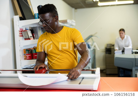 Focused middle-aged African American man cutting the printed sheet on the cutter in the typography 106156158