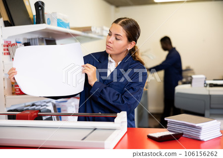 Attentive young female in a blue uniform verifying printed sheet during work in the printer shop Attentive young female in a blue uniform verifying printed sheet during work in the printer shop 106156262
