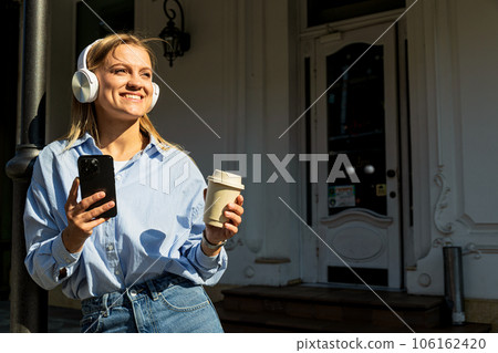 Smiling young woman in headphones with smartphone and paper cup in hands outdoors 106162420
