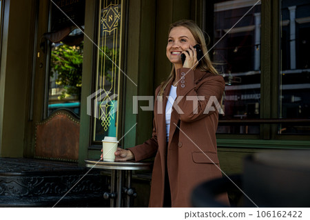 Smiling business woman with paper cup in hand talking on phone outdoors 106162422