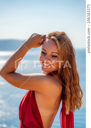 Smiling young woman in a red dress looks at the camera. A beautiful tanned girl enjoys her summer holidays at the sea. Portrait of a stylish carefree woman laughing at the ocean. 106163375
