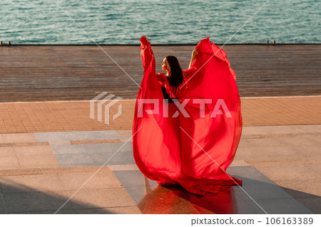 Sunrise red dress. A woman in a long red dress against the backdrop of sunrise, bright golden light of the sun's rays. The concept of femininity, harmony. 106163389