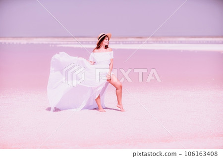 Woman in pink salt lake. She in a white dress and hat enjoys the scenic view of a pink salt lake as she walks along the white, salty shore, creating a lasting memory. 106163408
