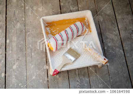 Paint roller and a brush in yellow tray liner on the background of old floorboard 106164598