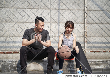 young asian adult male basketball player and female skateboarder sitting on court side talking chatting 106165541