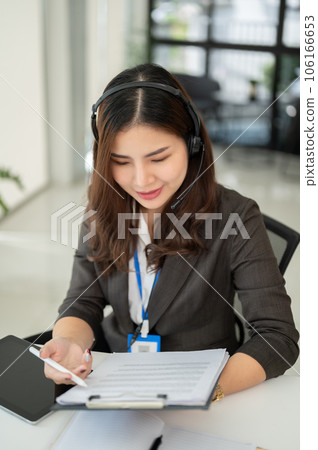 A beautiful Asian female call centre operator with a headset is examining report at her desk 106166653