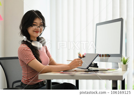 Portrait of young designer sitting at graphic studio in front of blank computer monitor and smiling at camera Portrait of young designer sitting at graphic studio in front of blank computer monitor and smiling at camera 106169125