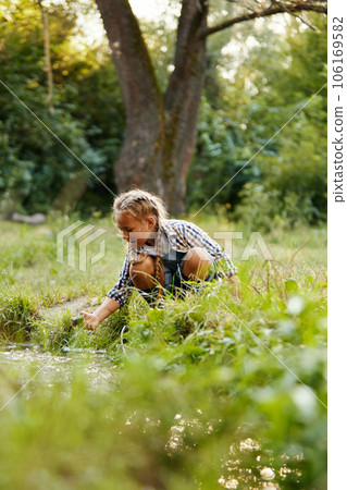 Little girl, child in casual clothes sitting near small river in forest on warm day. Nature exploration 106169582