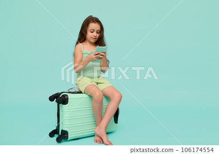 Little child girl using smartphone, sitting on suitcase, awaiting flight, checking mobile app, isolated blue background 106174554