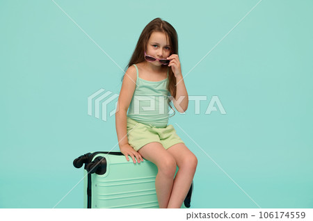 Conceptual portrait of a kid girl sitting on her suitcase, looking at camera, isolated over turquoise blue background 106174559