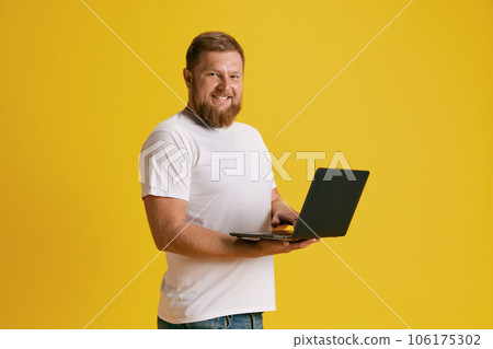 Smiling, positive, bearded man standing with laptop, working, shopping against yellow studio background Smiling, positive, bearded man standing with laptop, working, shopping against yellow studio background 106175302