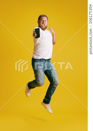 Man emotionally jumping with mobile phone against yellow studio background. Success, winning 106175304