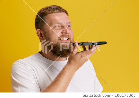 Bearded smiling man in casual white t-shirt talking mobile phone, recording voice message against yellow studio background 106175305