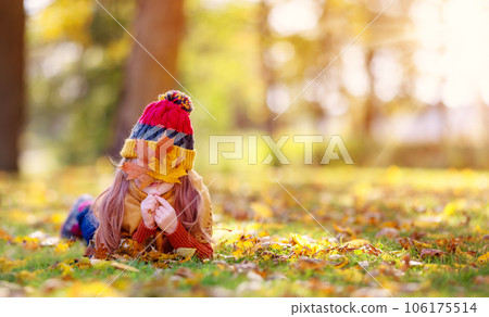 Playful little girl hiding her face behind autumnal leaf in the park. 106175514