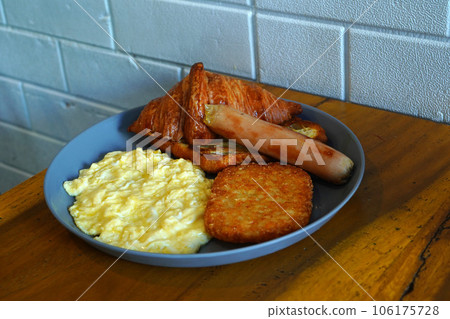 Breakfast consisting of hash browns, croissants, sausages, and scrambled eggs is served on gray ceramic plates placed on a wooden table. Breakfast consisting of hash browns, croissants, sausages, and scrambled eggs is served on gray ceramic plates placed on a wooden table. 106175728