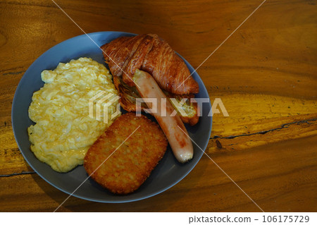 Breakfast consisting of hash browns, croissants, sausages, and scrambled eggs is served on gray ceramic plates placed on a wooden table. Breakfast consisting of hash browns, croissants, sausages, and scrambled eggs is served on gray ceramic plates placed on a wooden table. 106175729