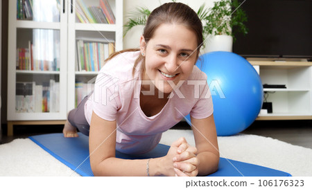 Portrait of smiling woman looking in camera while standing doing planking on fitness mat at home Portrait of smiling woman looking in camera while standing doing planking on fitness mat at home 106176323