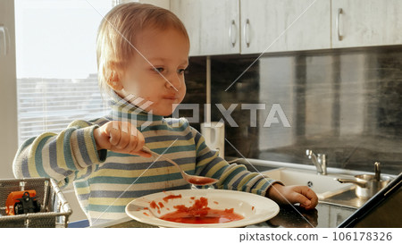 Cute messy boy eating soup with spoon and watching cartoons. Domestic food, gadget addiction, children healthy nutrition. Cute messy boy eating soup with spoon and watching cartoons. Domestic food, gadget addiction, children healthy nutrition. 106178326