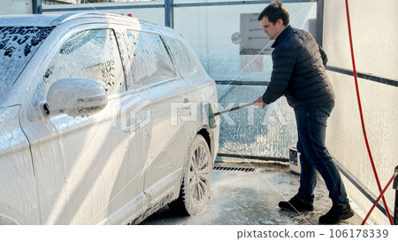 Young man washing his car with foam and brush at carwash. Automobile care, transport cleaning, dirty car. 106178339