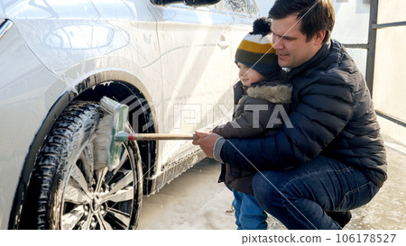 Smiling man with his baby son washing dirty car wheels at carwash. Automobile care, transport cleaning, dirty car. Smiling man with his baby son washing dirty car wheels at carwash. Automobile care, transport cleaning, dirty car. 106178527