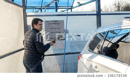 Young man washing his car with water jet at self service carwash. Automobile care, transport cleaning, dirty car. 106178536