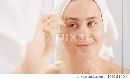 Smiling young woman wearing white towel plucking her eyebrows with cosmetic tweezers. Concept of beautiful female, makeup at home, skin care and domestic beauty industry. Smiling young woman wearing white towel plucking her eyebrows with cosmetic tweezers. Concept of beautiful female, makeup at home, skin care and domestic beauty industry. 106181404
