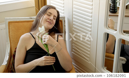 Elegant woman with white tulip sitting in armchair at big window. Female beauty, romantic mood, flowers at home. 106182091