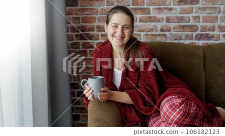 Happy smiling woman in pajamas covering with plaid and drinking tea on sofa. Female resting at home, lifestyle and relaxation. 106182123