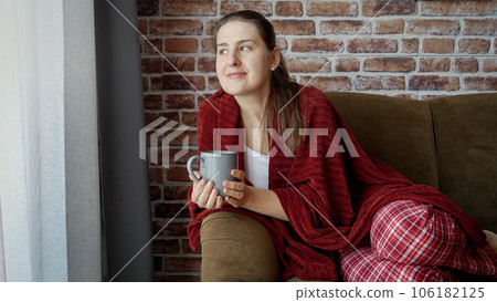 Smiling woman drinking tea under plaid and looking out of the window. Female resting at home, lifestyle and relaxation. 106182125